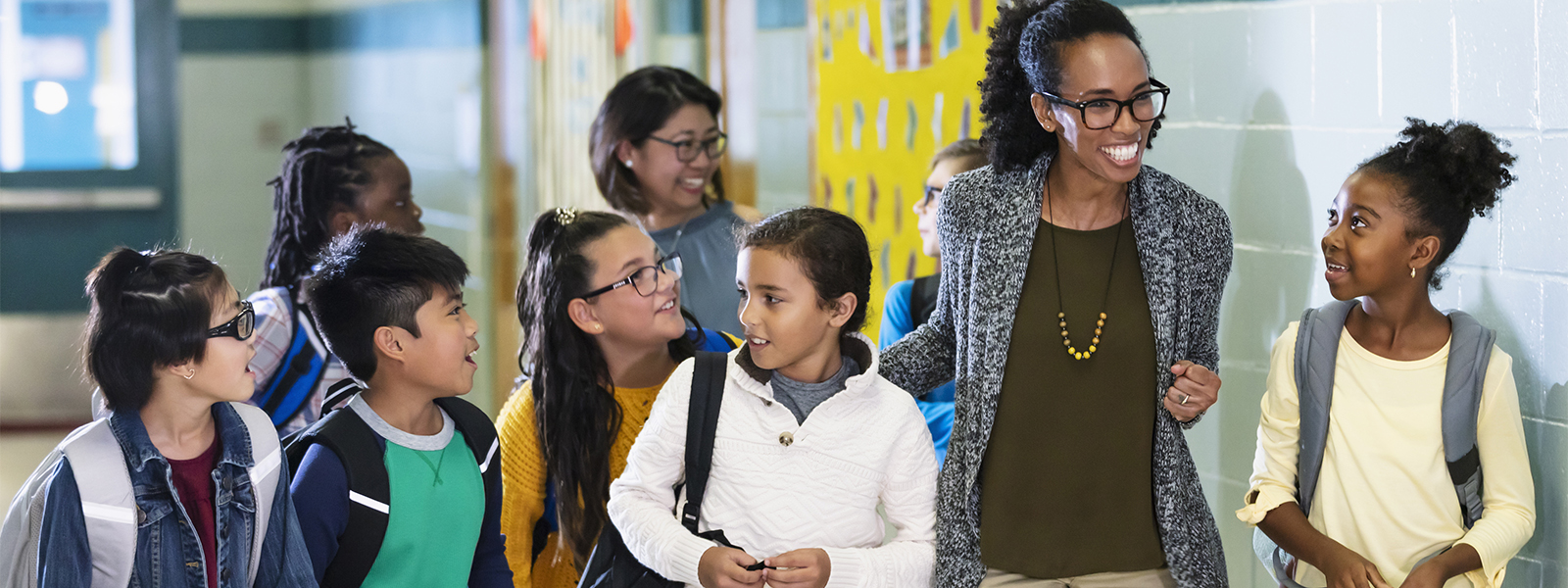 A multiracial group of elementary school children and their teachers walking and talking in a school hallway.